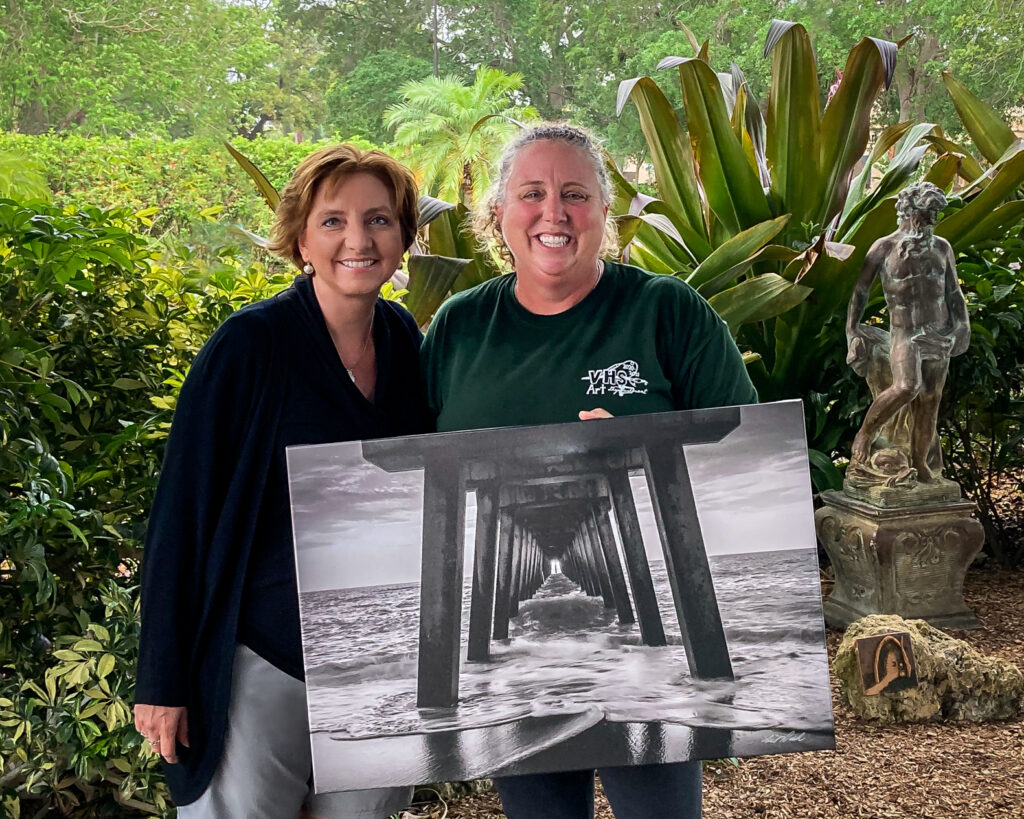 Portrait of Liesl Walsh and a client who purchased a print from her. The client is holding a canvas print of a black and white photo of the Venice Fishing Pier, Florida.