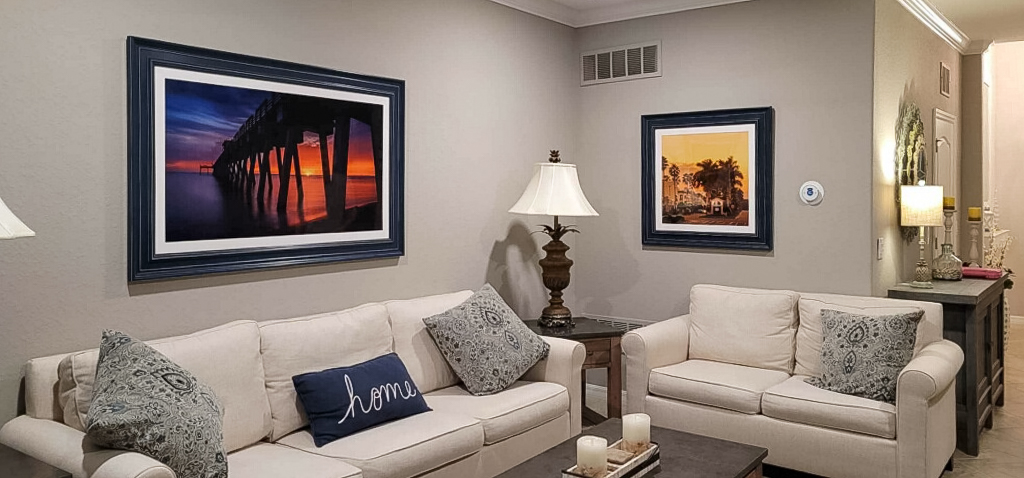 A living room with white couches and two prints hanging on the white walls. Each print is framed with a blue frame. The print on the left is the Venice Fishing Pier at sunset and the smaller print is downtown Venice, Florida with a yellow sunset sky.