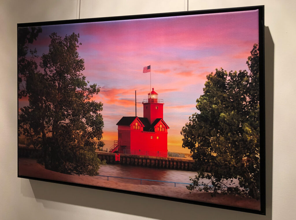 24 x 16 Canvas print of the Big Red Lighthouse in Holland, Michigan at sunset with a pink sky, with a tree on each side. The canvas is framed with a black floater frame and hangs on a white wall.