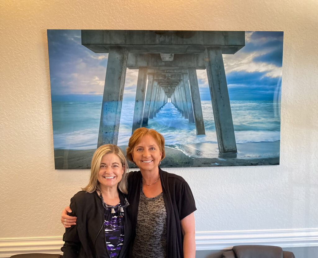 Portrait of Liesl Walsh and a local female dentist standing in front of a 60 x 34 metal print of the Venice Fishing Pier, Florida hanging on a white wall. The print has shades of blue, grey, and aqua.