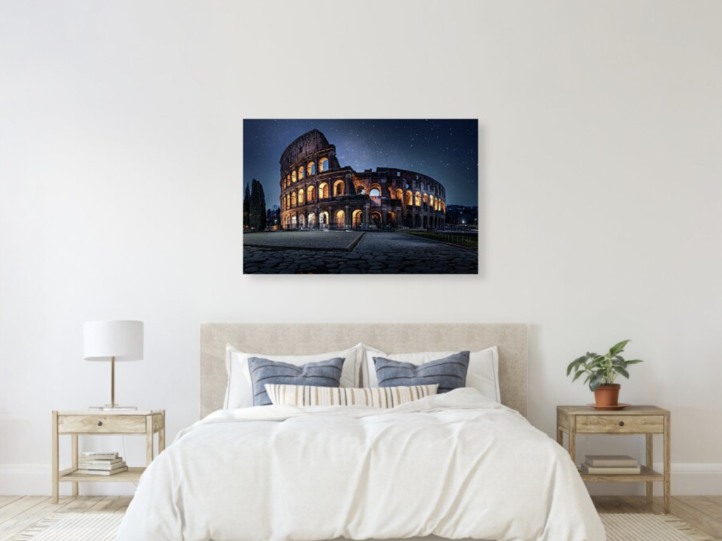 A bedroom mockup from Fine Art America with a print of the Colosseum in Rome at night hanging above a bed on a white wall. The bed has neutral colors and blue pillows.